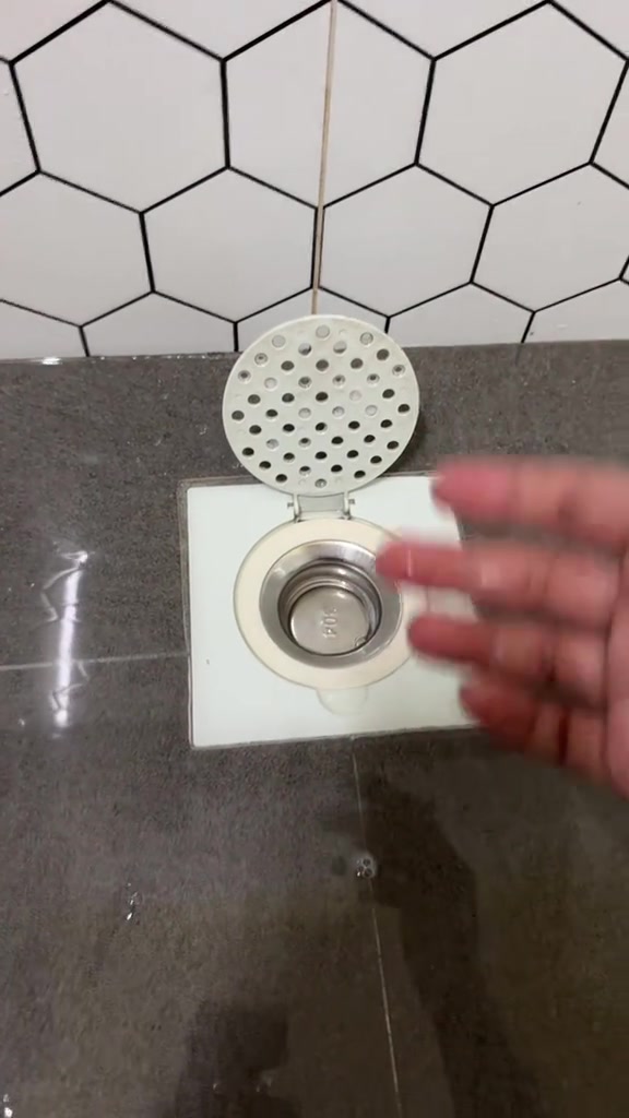 Core stainless steel bertanda 304 dalam floor trap segi empat putih dengan penutup bulat berlubang berengsel terbuka di hadapan dinding jubin heksagon