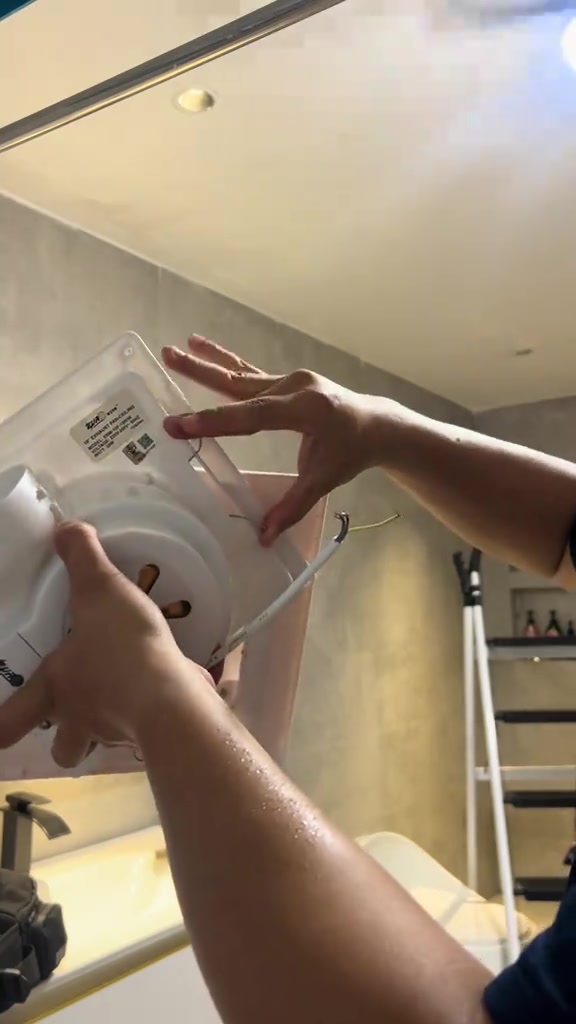 Tangan memegang unit exhaust fan dari bawah menunjukkan label spesifikasi dan wayar sambungan, di bawah siling bilik air
