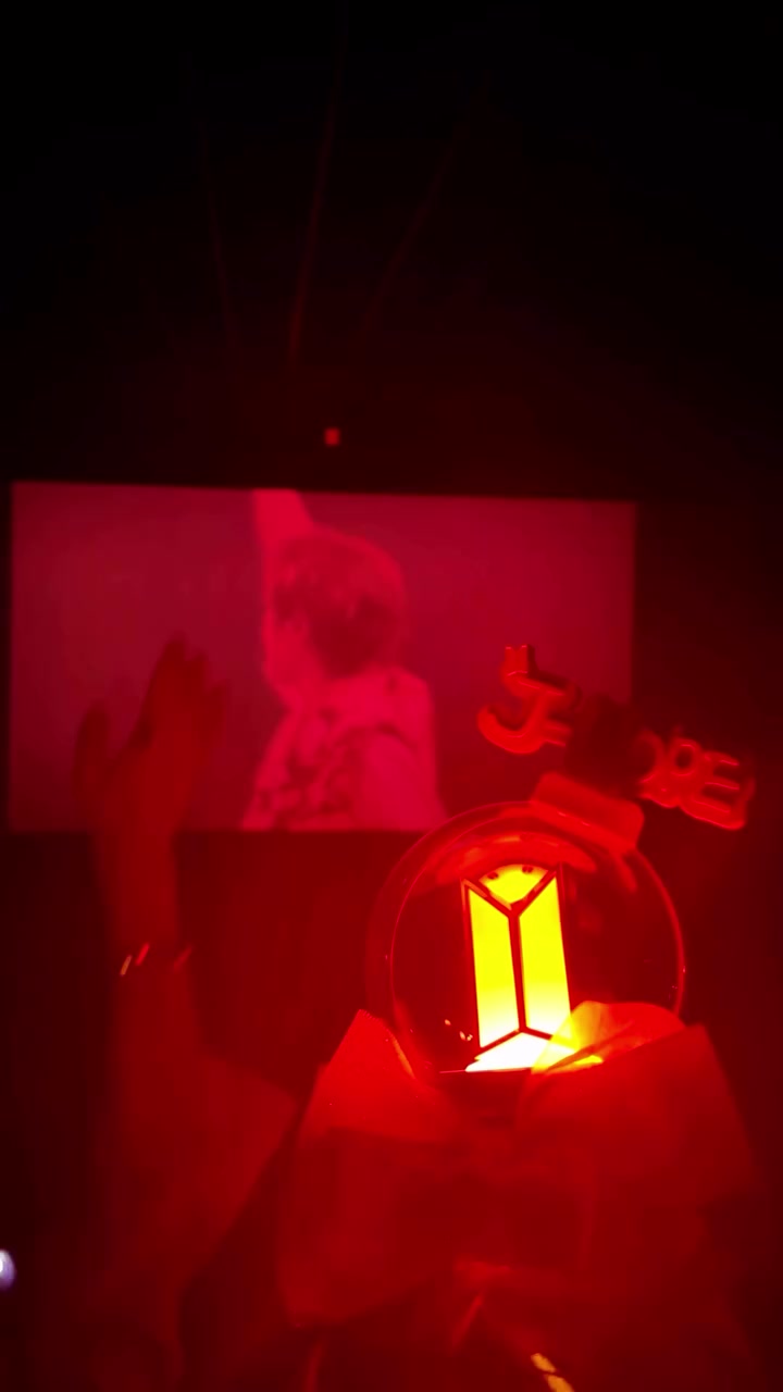 Lightstick ARMY bomb phát sáng màu vàng gắn nơ trang trí được giơ lên trong phòng chiếu nhuốm ánh đỏ, phía sau là màn hình rạp chiếu bóng nghệ sĩ từ phía sau