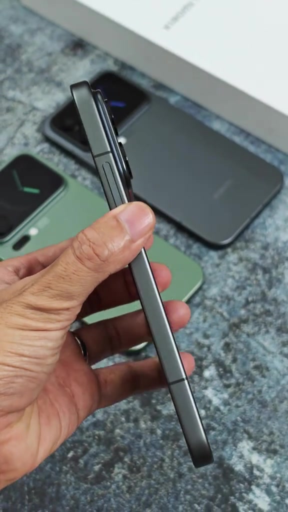 Cạnh sườn màu xám của Xiaomi 17 Pro Max với khung kim loại phẳng, cầm trên tay, phía sau là một máy xanh và một máy xám đặt trên thảm