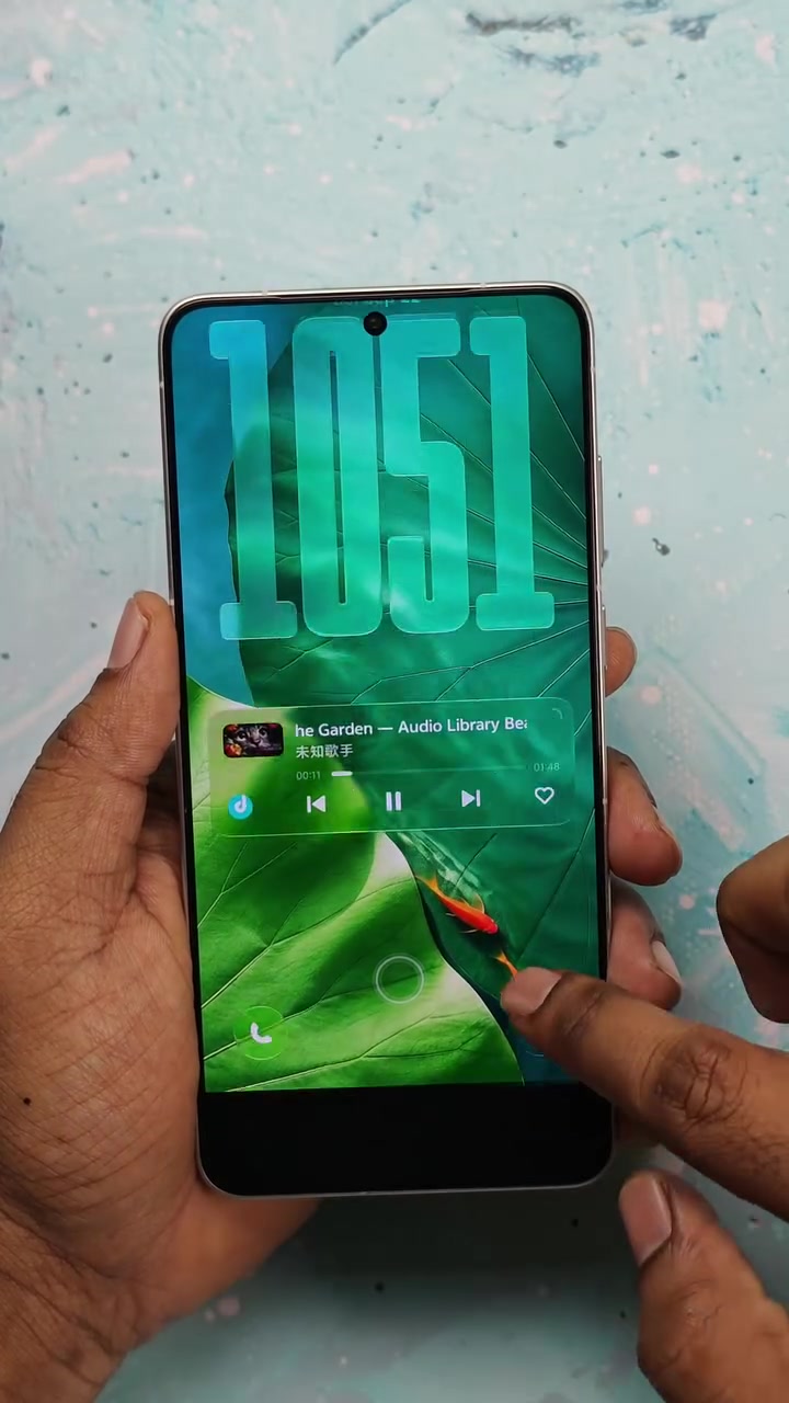 Điện thoại vivo chạy OriginOS 7 với màn khóa đồng hồ số cỡ lớn trên nền lá xanh và widget nhạc hiển thị chữ Trung