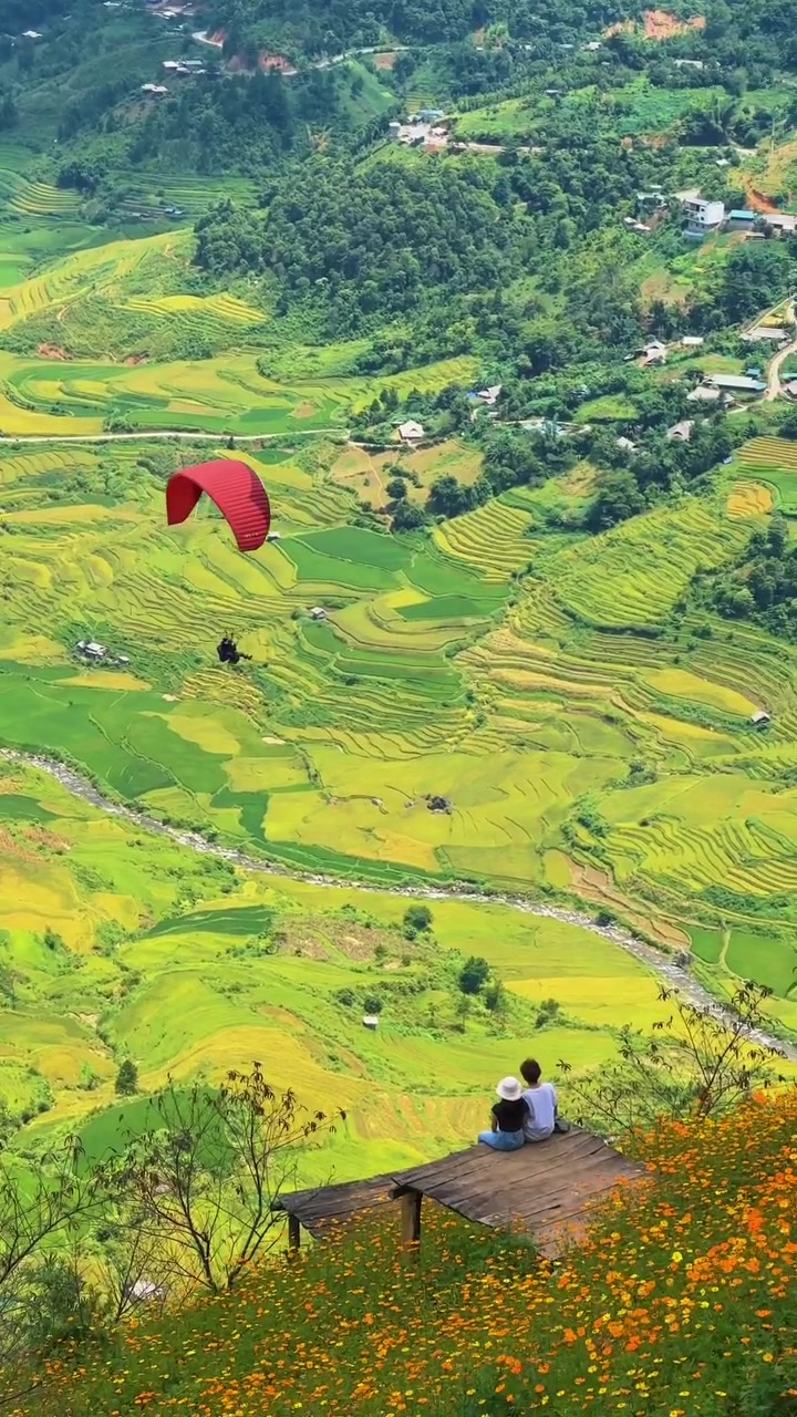 Hai người ngồi sát nhau nhìn từ phía sau trên sàn gỗ giữa đồi hoa, cùng ngắm thung lũng ruộng bậc thang, xa xa một cánh dù lượn màu đỏ