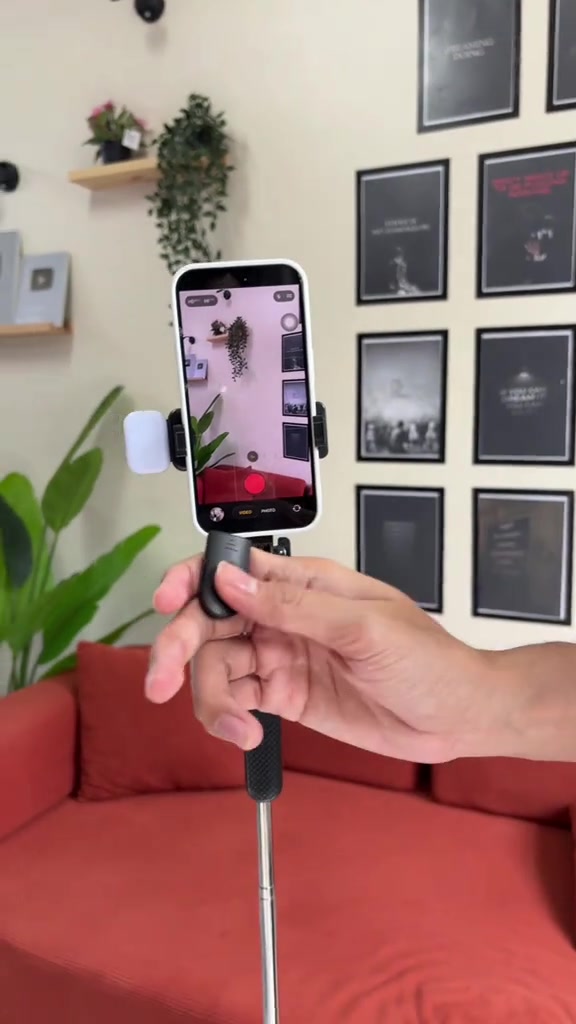 Tangan memegang remote Bluetooth kecil di hadapan telefon yang dipasang pada tripod selfie stick