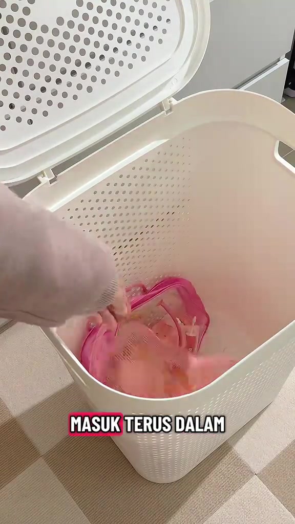Tangan memasukkan beg mesh merah jambu berisi pakaian ke dalam bakul plastik putih berlubang
