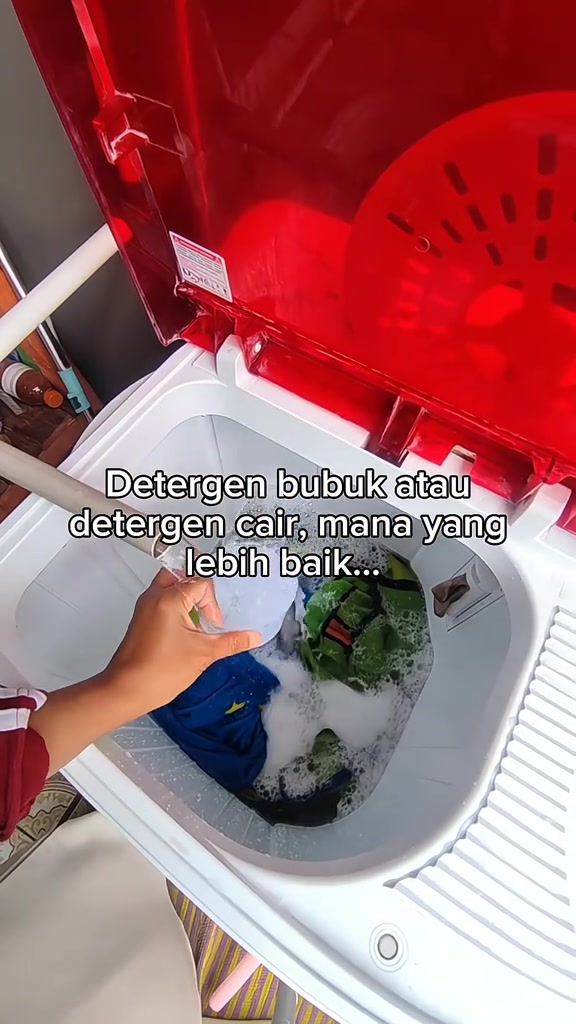 Tangan memegang penutup sukatan di atas air berbuih dalam mesin basuh top load yang sedang membasuh pakaian berwarna