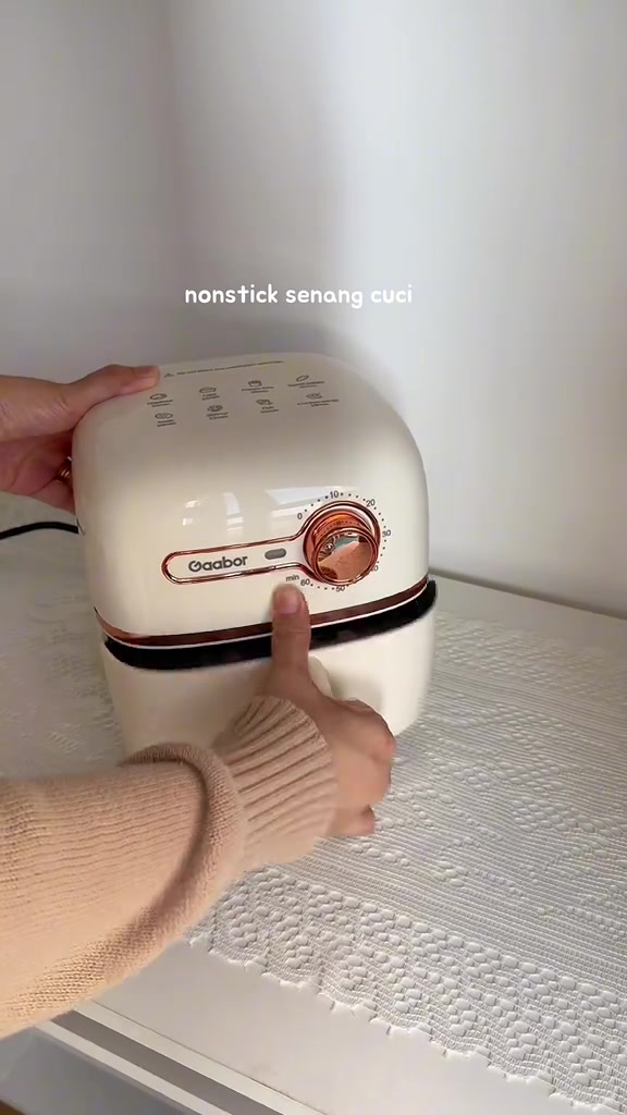 Air fryer mini krim Gaabor atas meja putih dengan kabel kuasa di sisi, tangan menunjuk ke badan dia