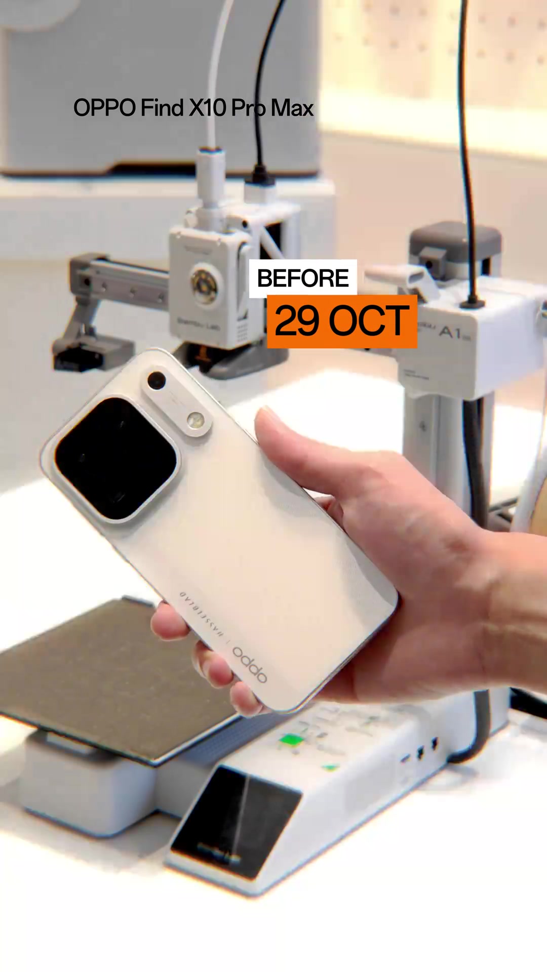 Tangan memegang OPPO Find X10 Pro Max warna putih di hadapan pencetak 3D Bambu Lab A1 Mini di meja pameran