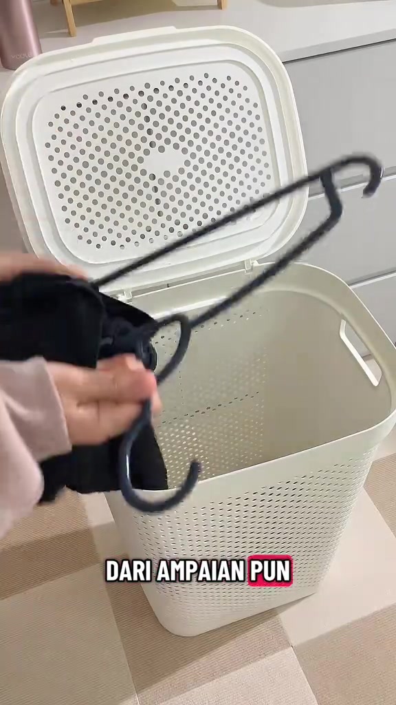 Tangan memasukkan baju hitam berhanger ke dalam bakul plastik putih berlubang dengan penutup terbuka