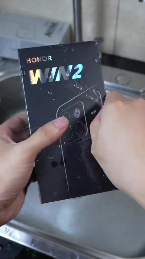 Kotak hitam HONOR WIN 2 dipegang dua tangan di atas sinki stainless