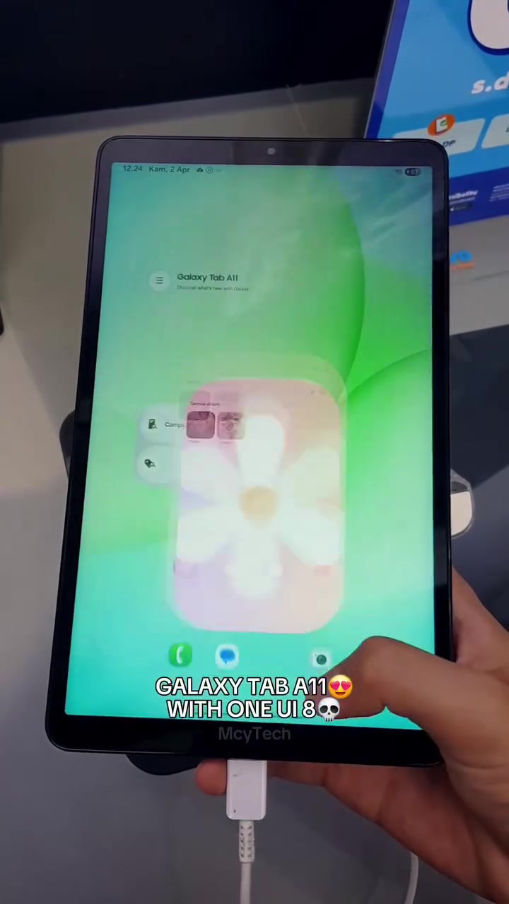 Samsung Galaxy Tab A11 dipegang satu tangan dengan skrin hijau One UI 8 memaparkan nama model, kabel pengecas terpasang di bawah