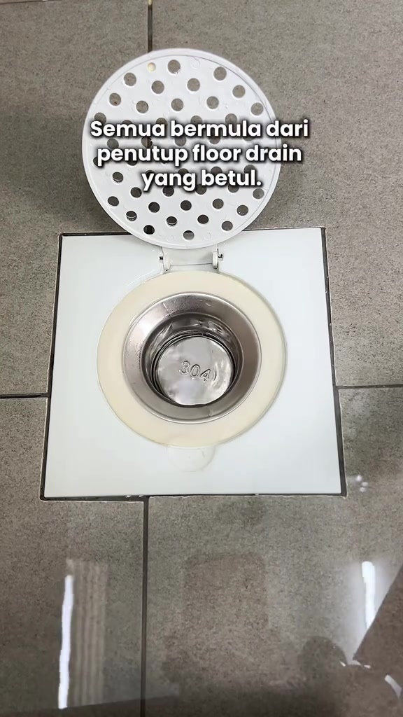 Pandangan atas floor trap segi empat putih dengan core stainless bertanda 304 dan penutup bulat berlubang berengsel terbuka di lantai jubin
