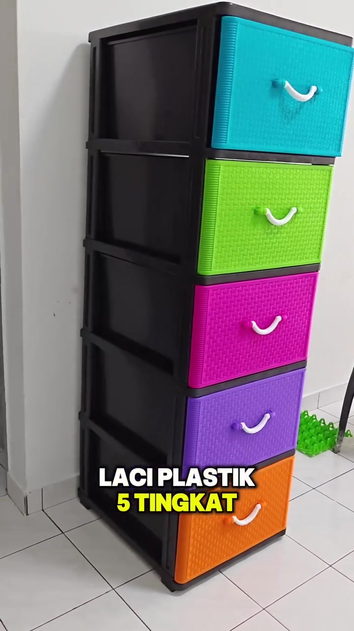 Almari laci plastik lima tingkat rangka hitam dengan laci biru, hijau, merah jambu, ungu dan oren berdiri di tepi dinding