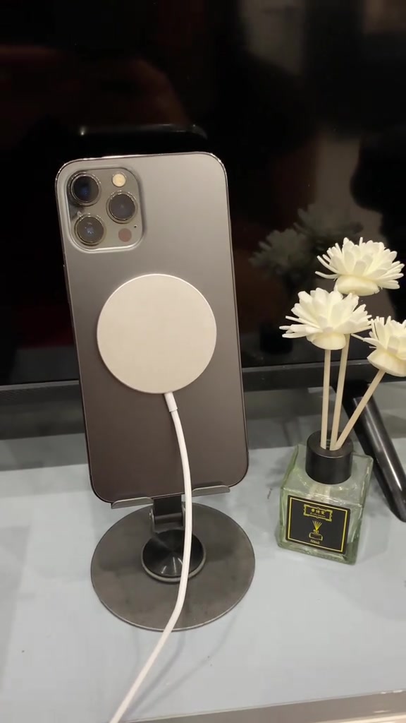 iPhone màu xám tựa trên giá đỡ, đĩa sạc MagSafe màu trắng hút giữa lưng máy, dây cáp thả xuống bàn