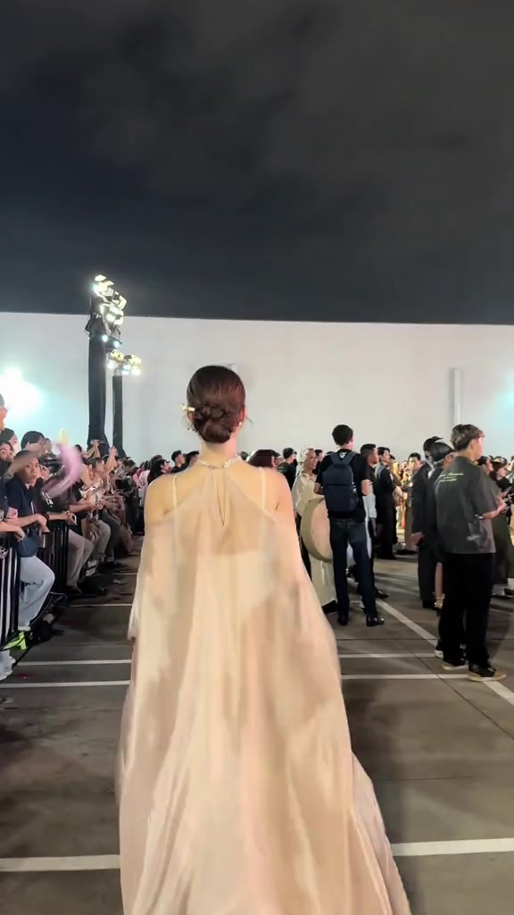 Một nghệ sĩ váy dạ hội màu champagne dáng cape nhìn từ phía sau, đi giữa lối đi đêm ra mắt; bên trái là hàng khán giả ngồi sau rào chắn giơ tay vẫy, phía xa là cột đèn sân khấu
