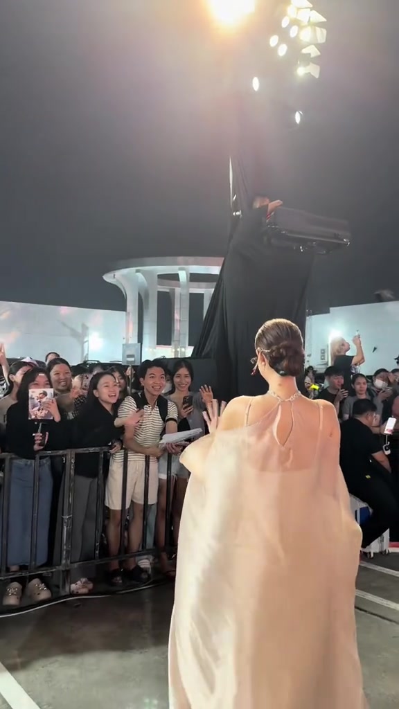 Nghệ sĩ váy cape màu champagne nhìn từ phía sau, vẫy tay về phía khu fan đứng sau rào chắn dưới dàn đèn sân khấu đêm sự kiện