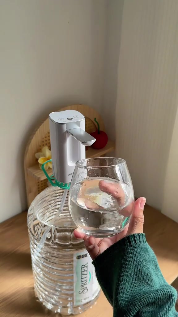 Tangan memegang gelas berisi air di bawah muncung pam botol elektrik