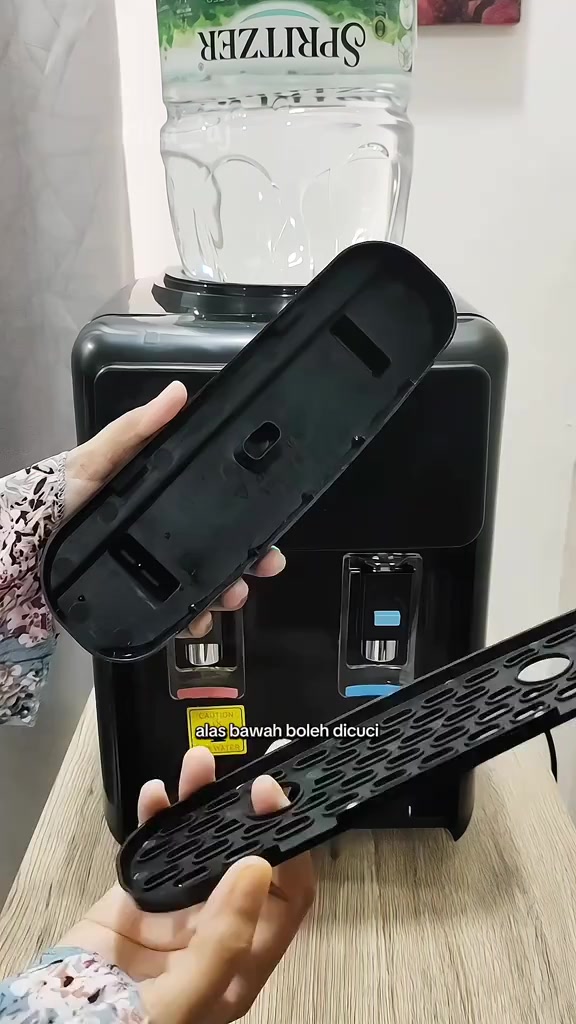 Dispenser tabletop hitam dengan botol air di atas, tangan memegang alas bawah yang boleh tanggal untuk dicuci