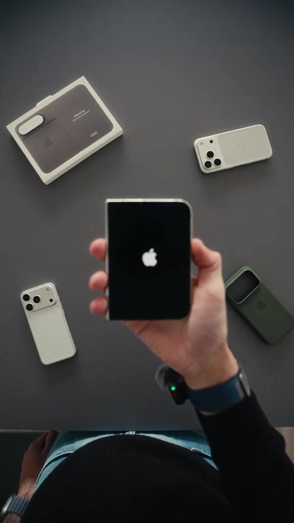 iPhone Duo tertutup dengan logo Apple dipegang di tangan, dikelilingi dua telefon iPhone lain dan kotak di atas meja