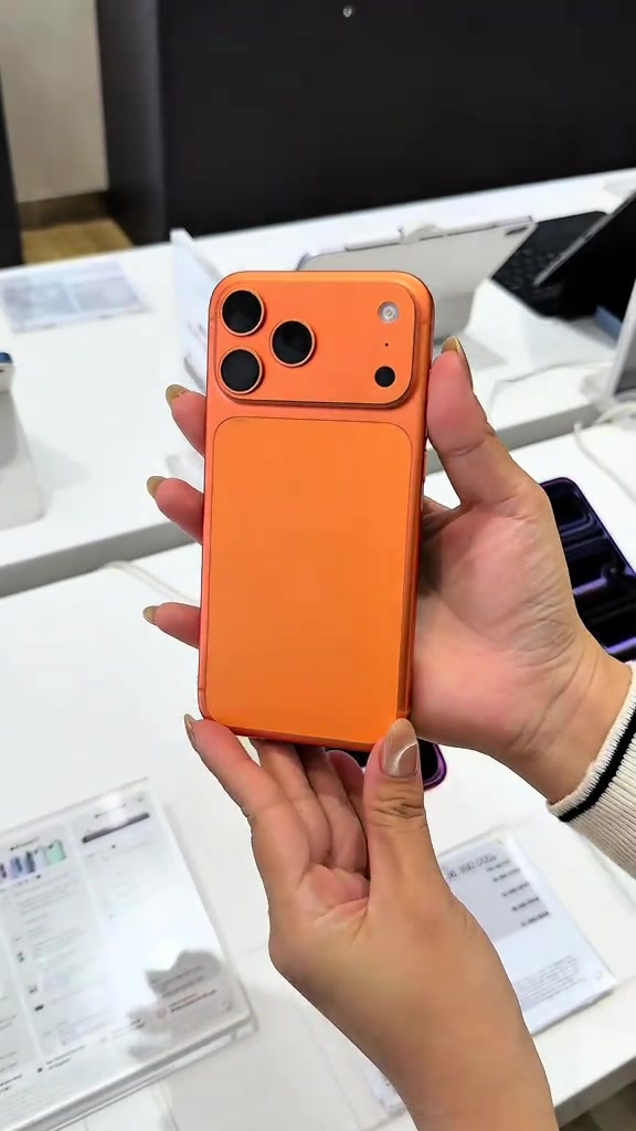 Hai tay cầm một chiếc điện thoại màu cam có cụm ba camera, nền là bàn trải nghiệm màu trắng trong cửa hàng