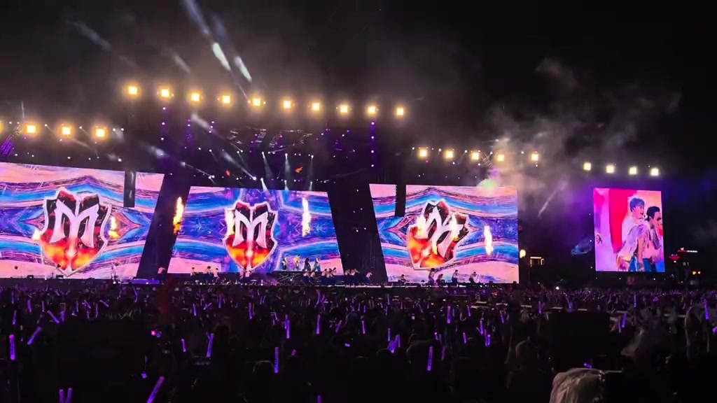 Toàn cảnh sân khấu concert ban đêm với màn LED lớn hiện logo chữ M bốc lửa, dàn đèn tím và biển lightstick tím phía khán đài