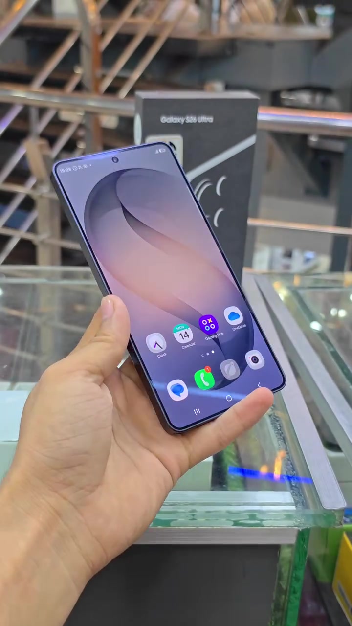 Galaxy S26 Ultra dengan skrin hidup dipegang satu tangan, kotak asal berdiri di latar belakang