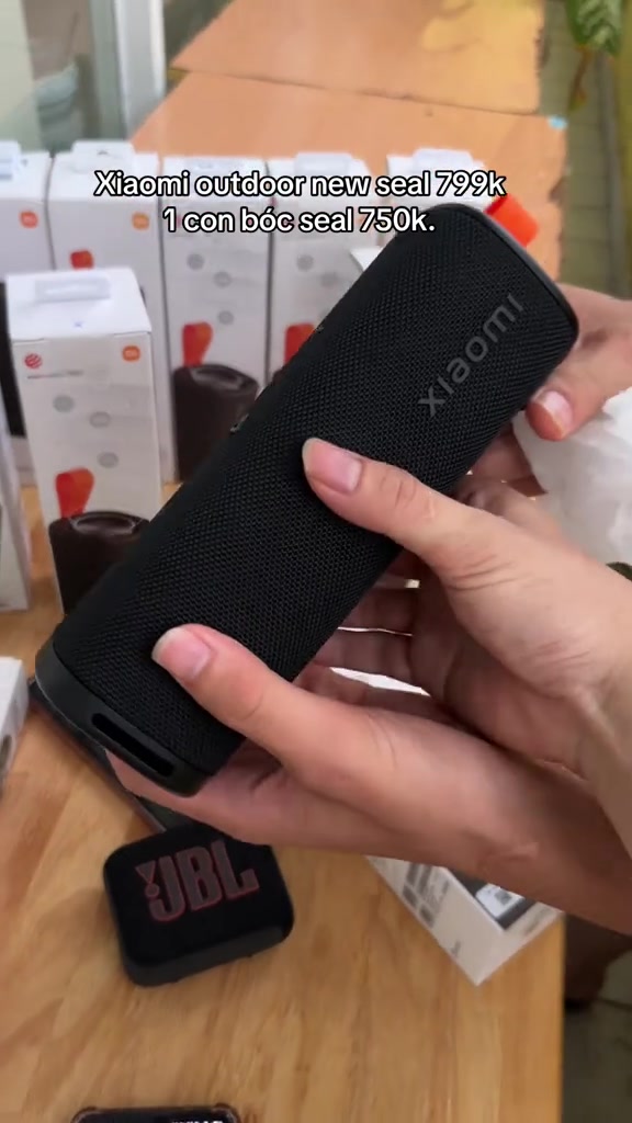Hai tay cầm chiếc loa bluetooth Xiaomi Sound Outdoor màu đen dạng trụ bọc vải, phía sau là các hộp sản phẩm