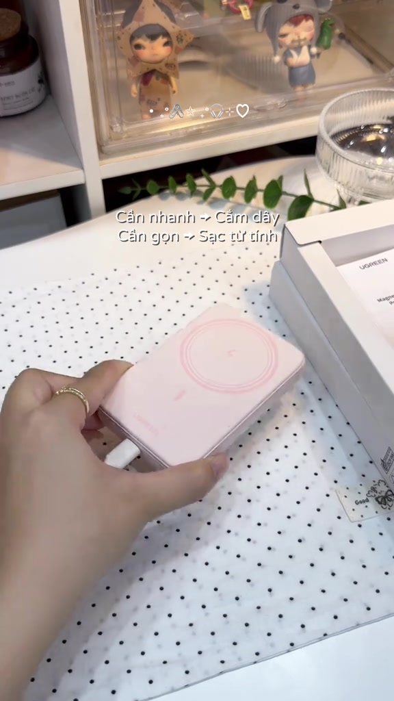 Cục sạc dự phòng từ tính Ugreen màu hồng nằm trong lòng bàn tay, cáp trắng cắm vào cạnh bên, hộp sản phẩm phía sau