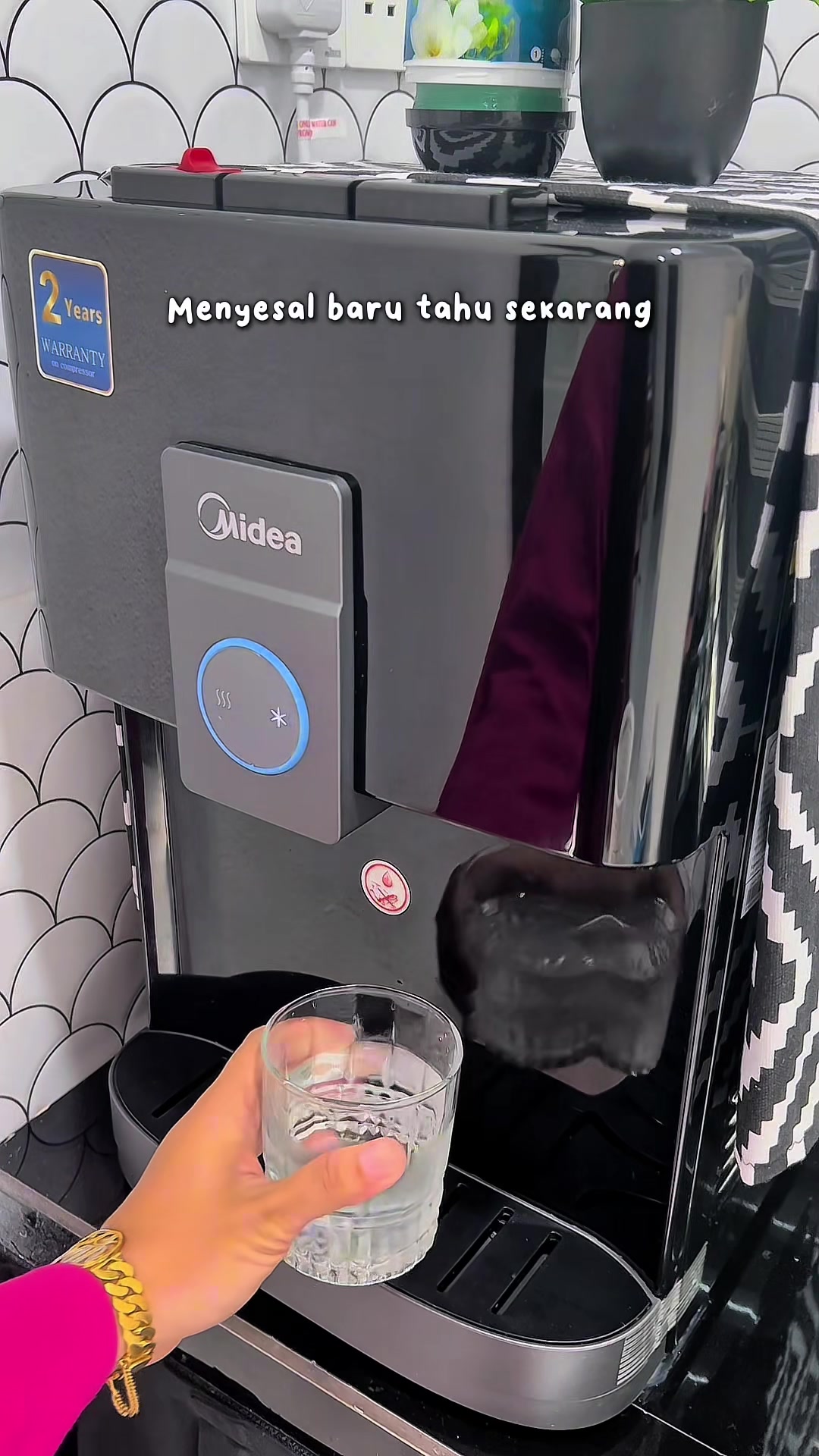 Tangan memegang gelas air yang diisi dari dispenser Midea di dapur