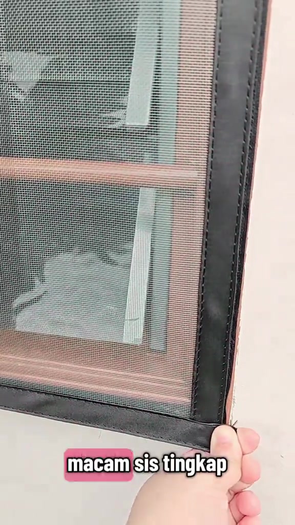 Tangan memegang penjuru helai jaring fiberglass kelabu dengan tepi jahitan hitam di hadapan tingkap