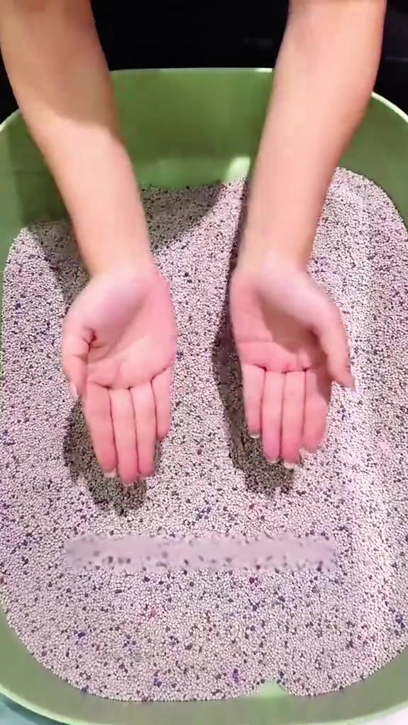 Sepasang tangan terbuka di atas pasir kucing dalam tray hijau dengan ketul air kelihatan di tengah