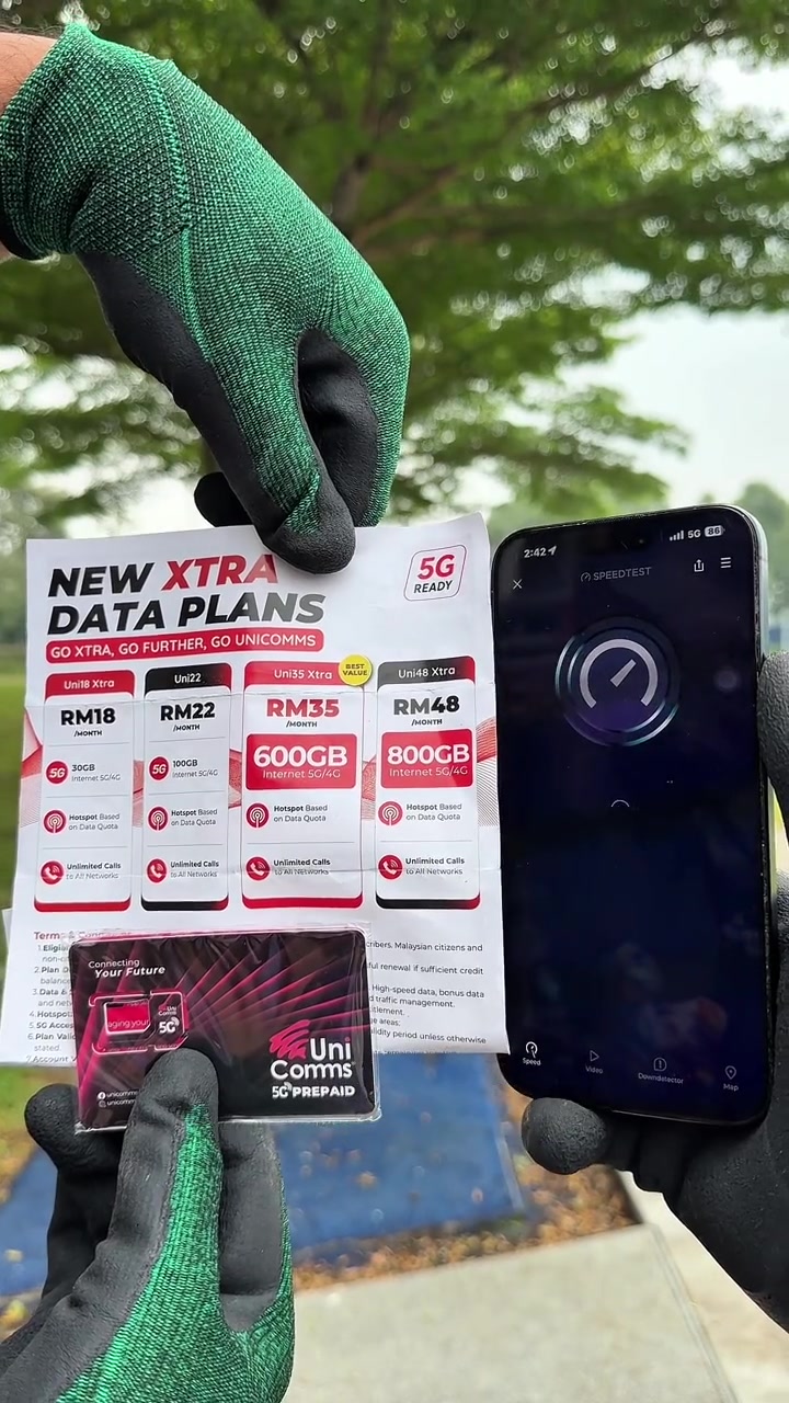 Tangan bersarung tangan hijau memegang helai senarai pelan UniComms Xtra dari RM18 hingga RM48 beserta pek SIM UniComms 5G Prepaid, di sebelah telefon yang sedang menjalankan ujian kelajuan