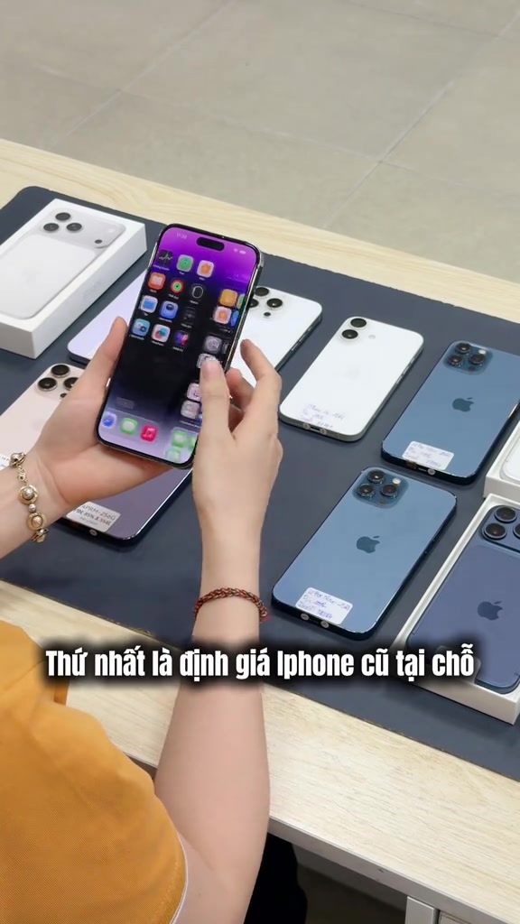 Bàn tay cầm một chiếc iPhone đang sáng màn hình, phía dưới là mặt bàn bày nhiều chiếc iPhone cũ úp lưng chờ định giá