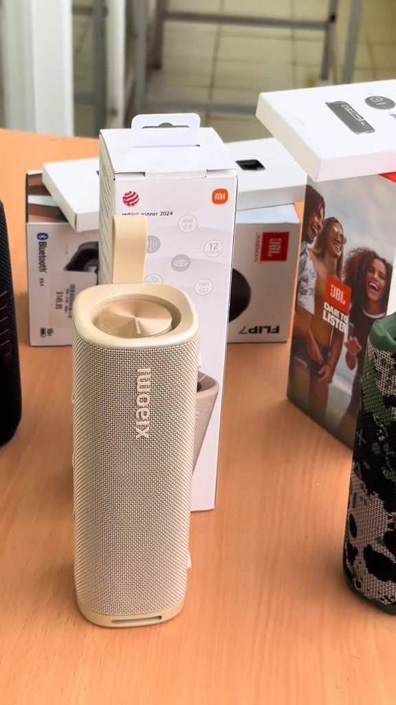 Loa Xiaomi Sound Outdoor bản màu kem đứng trên bàn, phía sau là hộp Xiaomi và hộp JBL Flip 7
