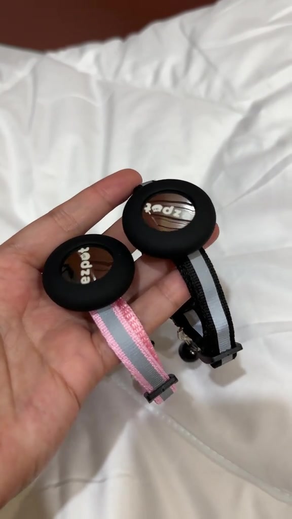 Tangan memegang dua tag penjejak bulat yang sudah dipasang pada collar kucing reflektif warna merah jambu dan hitam