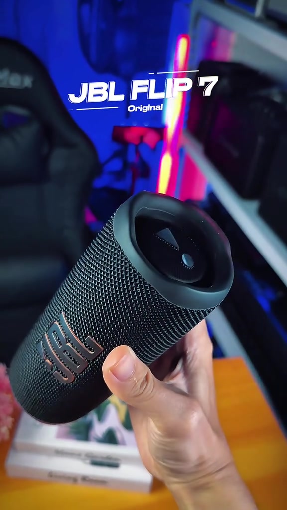 Tangan memegang speaker JBL Flip 7 hitam berbentuk silinder di depan rak barang gadget