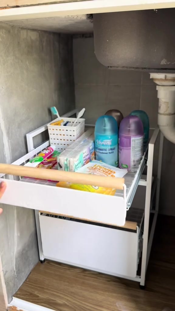 Rak drawer putih di bawah sinki berisi stok cleaning dan penyegar udara berdiri tegak, paip putih kelihatan di sebelah kanan