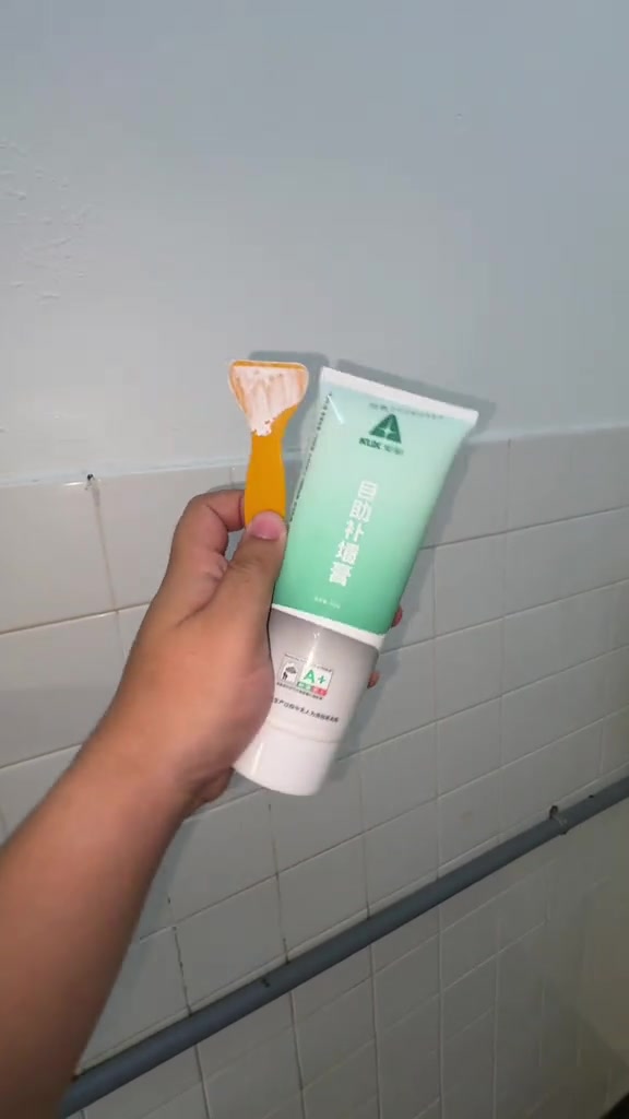 Tangan memegang tiub paste penampal dinding hijau muda bersama spatula oren berbaki paste putih