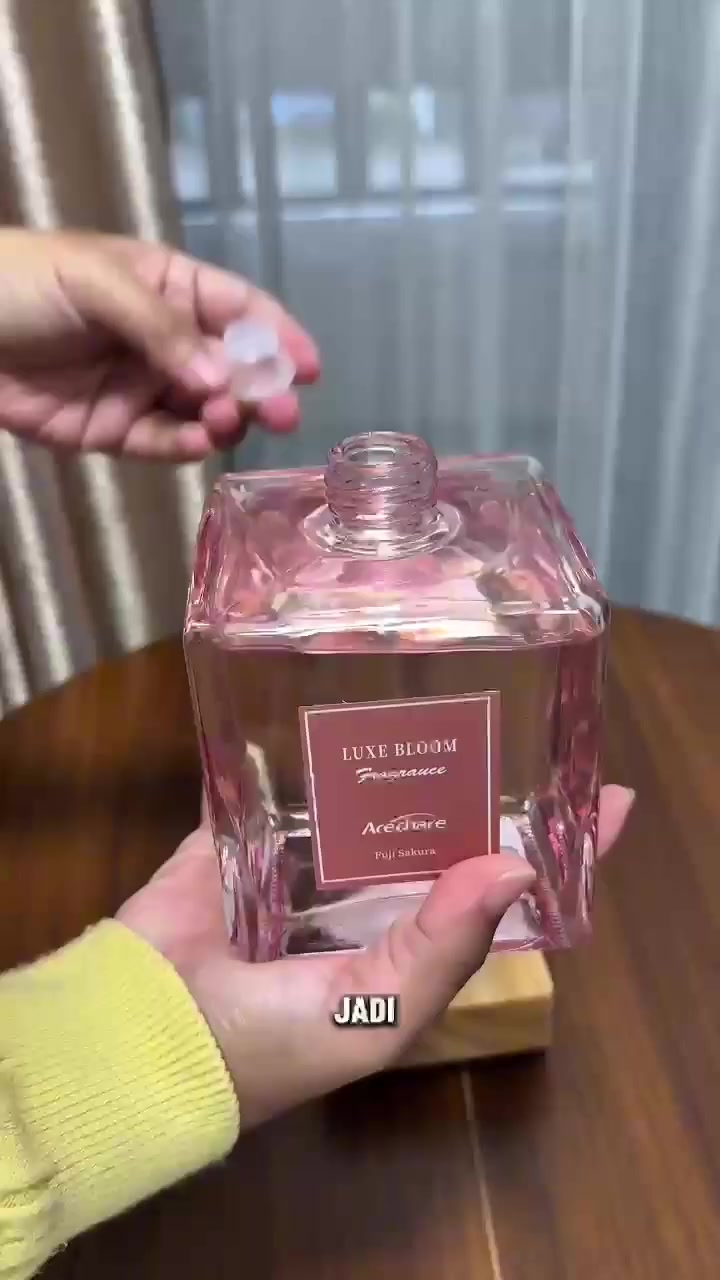Tangan memegang botol reed diffuser kaca merah jambu berlabel Luxe Bloom dengan penutup kayu