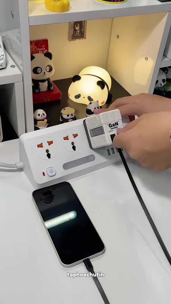 Củ sạc trắng in chữ GaN cắm trên ổ điện kéo dài trên bàn, một sợi cáp dẹt chạy xuống điện thoại đang sạc