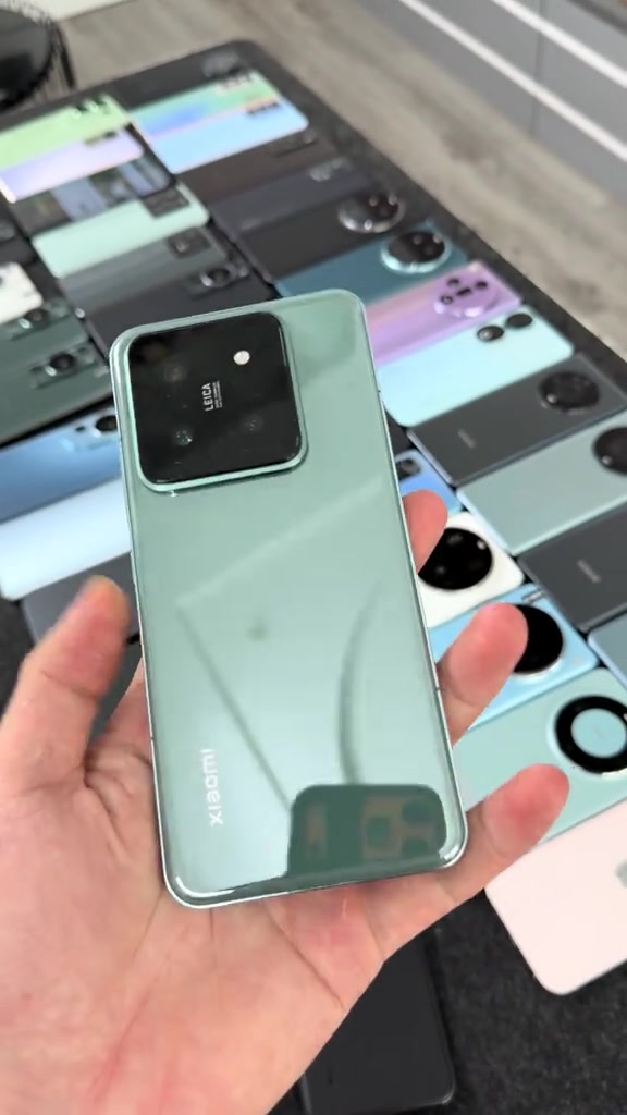 Một chiếc Xiaomi 14 Pro màu xanh nhạt cầm trên tay trước quầy bày hàng loạt điện thoại cũ đủ màu sắc