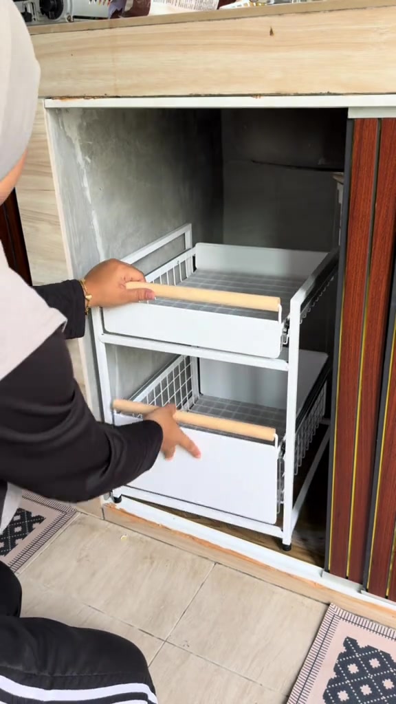 Rak drawer dua tingkat warna putih dimasukkan ke dalam ruang bawah sinki beton yang kosong oleh seorang wanita bertudung, nampak dari belakang