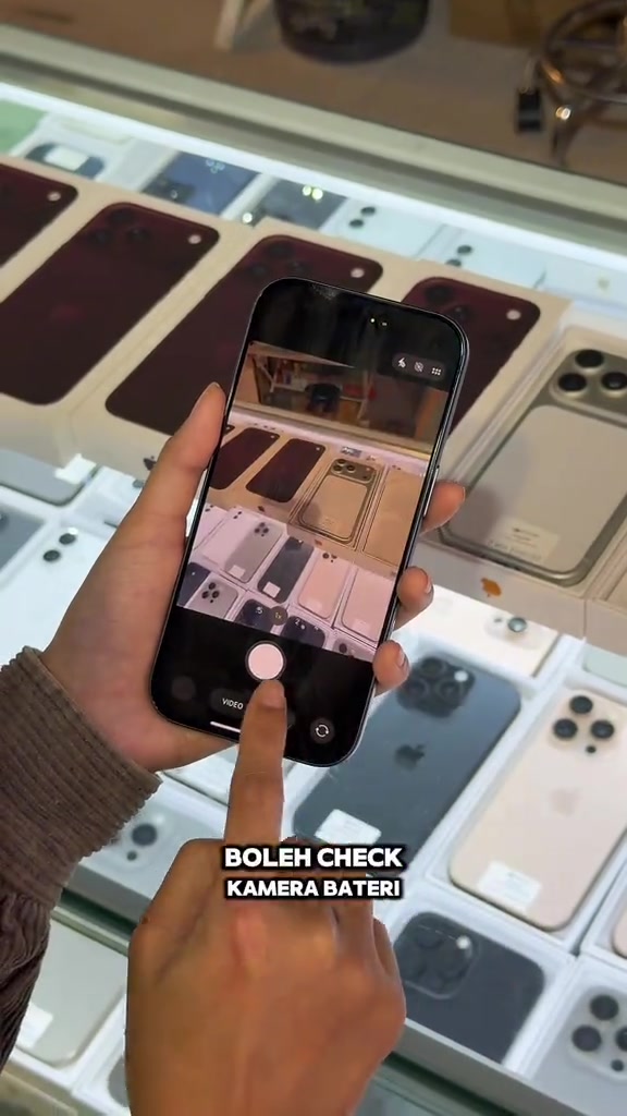 iPhone di tangan membuka aplikasi kamera untuk memeriksa kamera dan bateri di hadapan rak iPhone terpakai