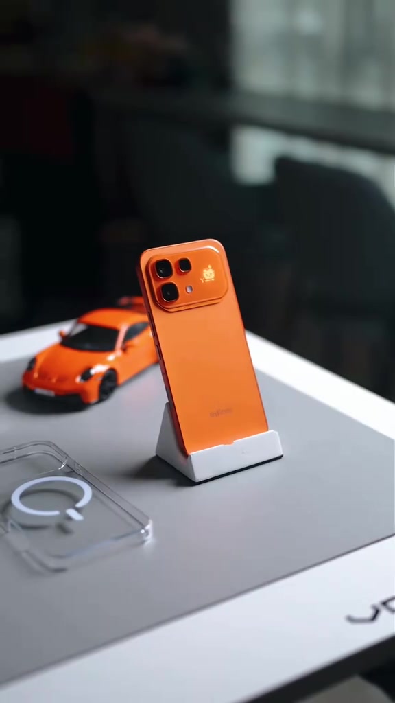 Infinix NOTE 60 Pro warna jingga berdiri pada stand putih di atas meja, sebelah kereta mainan jingga dan case lutsinar