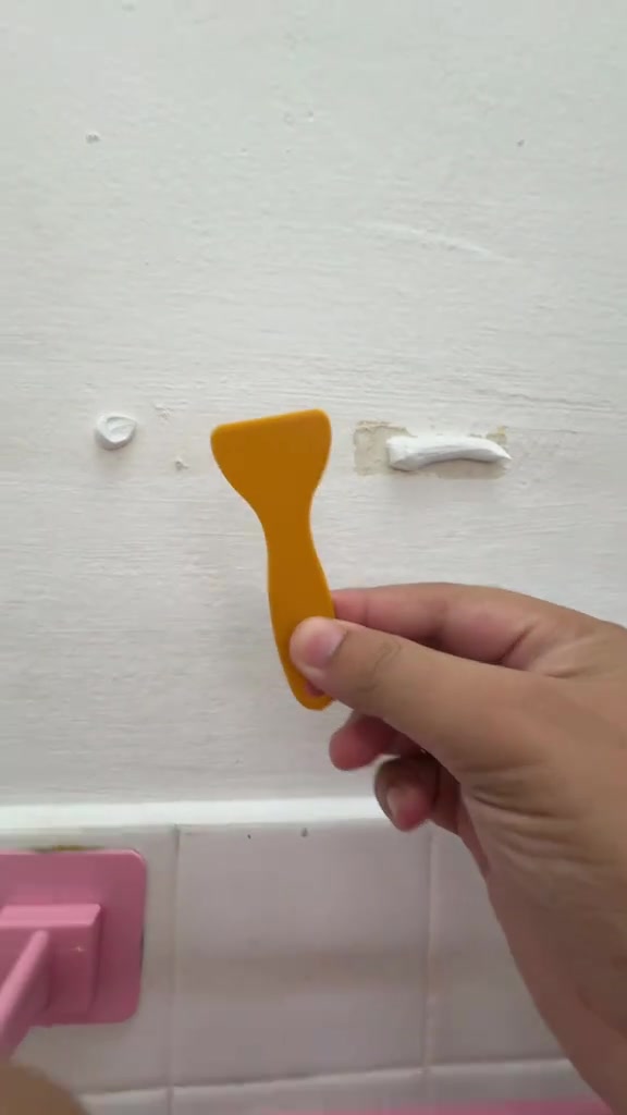Tangan memegang spatula plastik oren di depan dinding putih dengan tompok paste penampal pada lubang paku