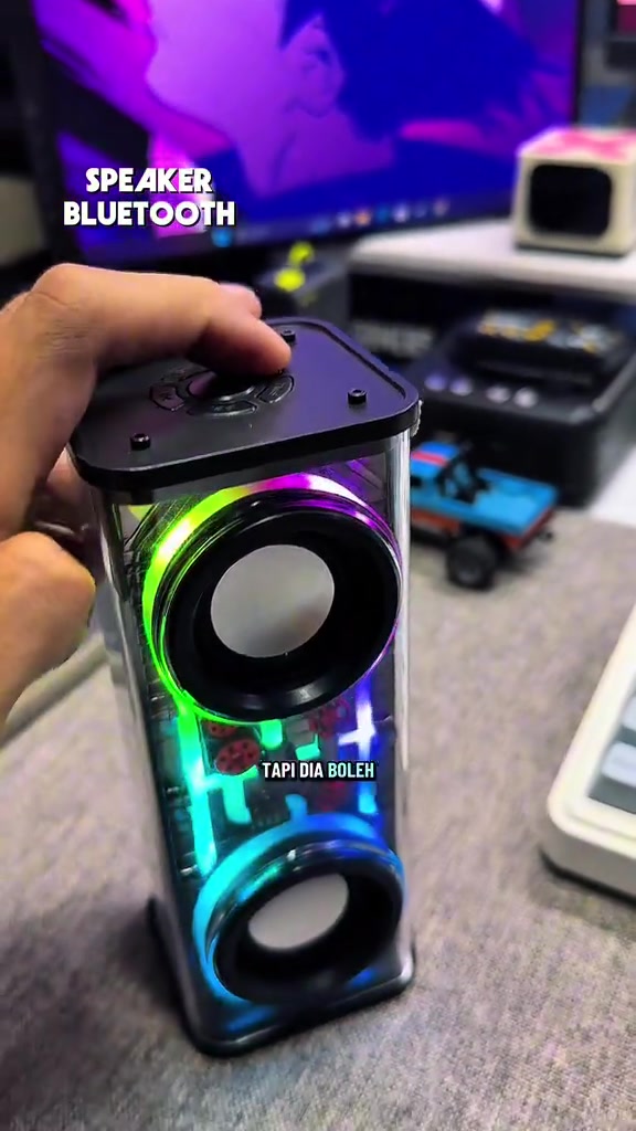 Tangan memegang speaker bluetooth kecil badan lutsinar dengan dua driver dan lampu RGB menyala