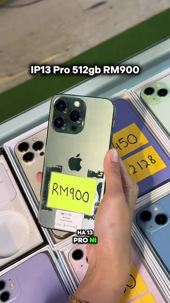 iPhone 13 Pro hijau di tangan dengan tag harga kuning RM900 di hadapan rak iPhone terpakai
