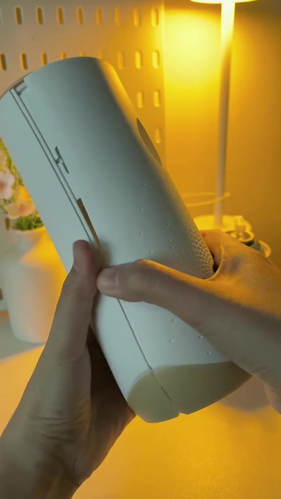 Tangan memegang dispenser pewangi automatik putih berbentuk silinder dalam bilik berlampu kuning