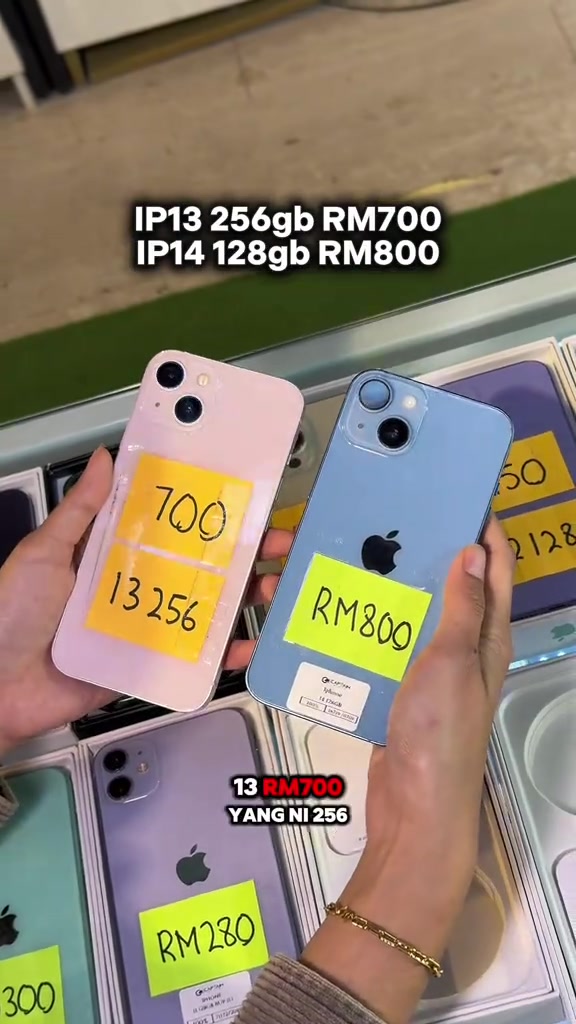 Dua iPhone terpakai di tangan dengan tag harga kuning RM700 (13 256) dan RM800 di hadapan rak kaca kedai
