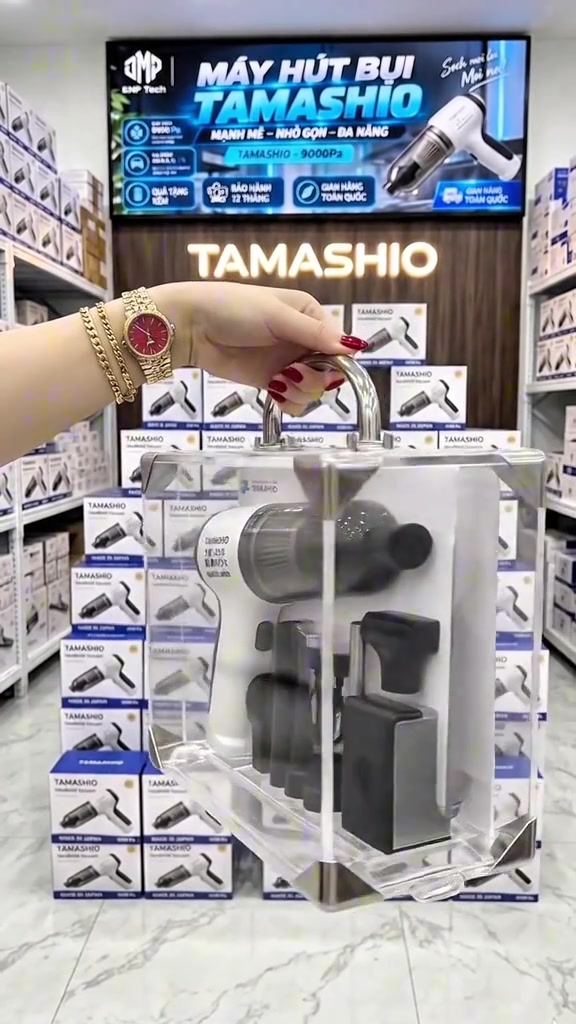 Tay xách hộp mica đựng máy hút bụi cầm tay và các đầu hút, phía sau là kệ xếp đầy hộp Tamashio và màn hình banner quảng cáo 9000Pa, bảo hành 12 tháng