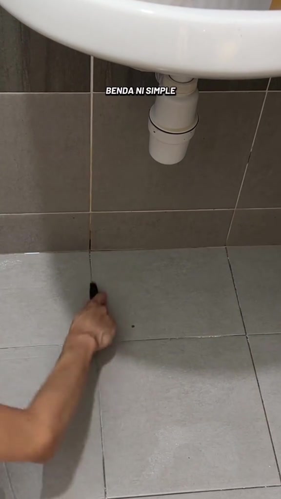 Tangan memegang alat kikis kecil hitam di celah nat jubin lantai bawah sinki bilik air